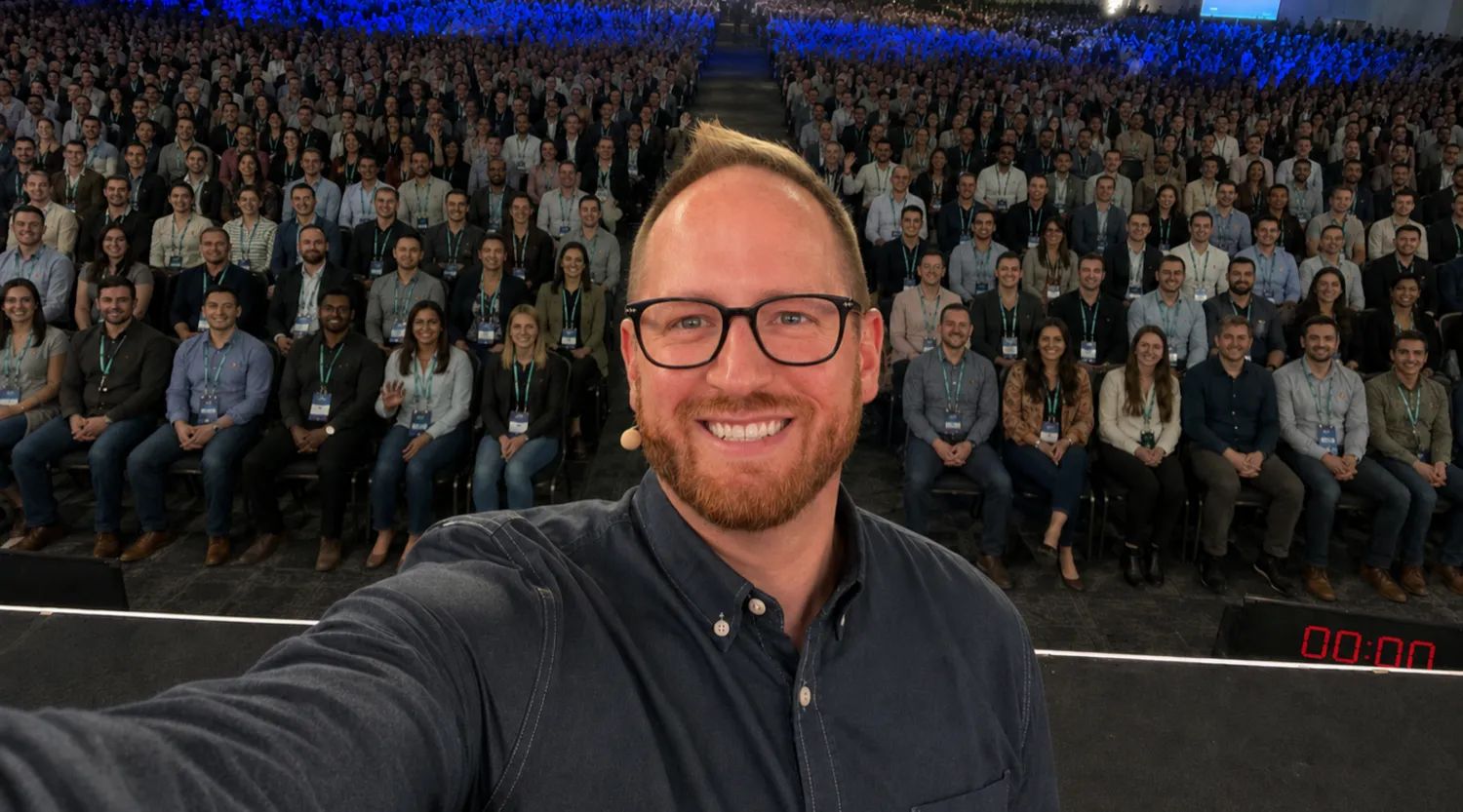 John Palmer on stage, taking a selfie with a full auditorium of several thousand people seated behind him.